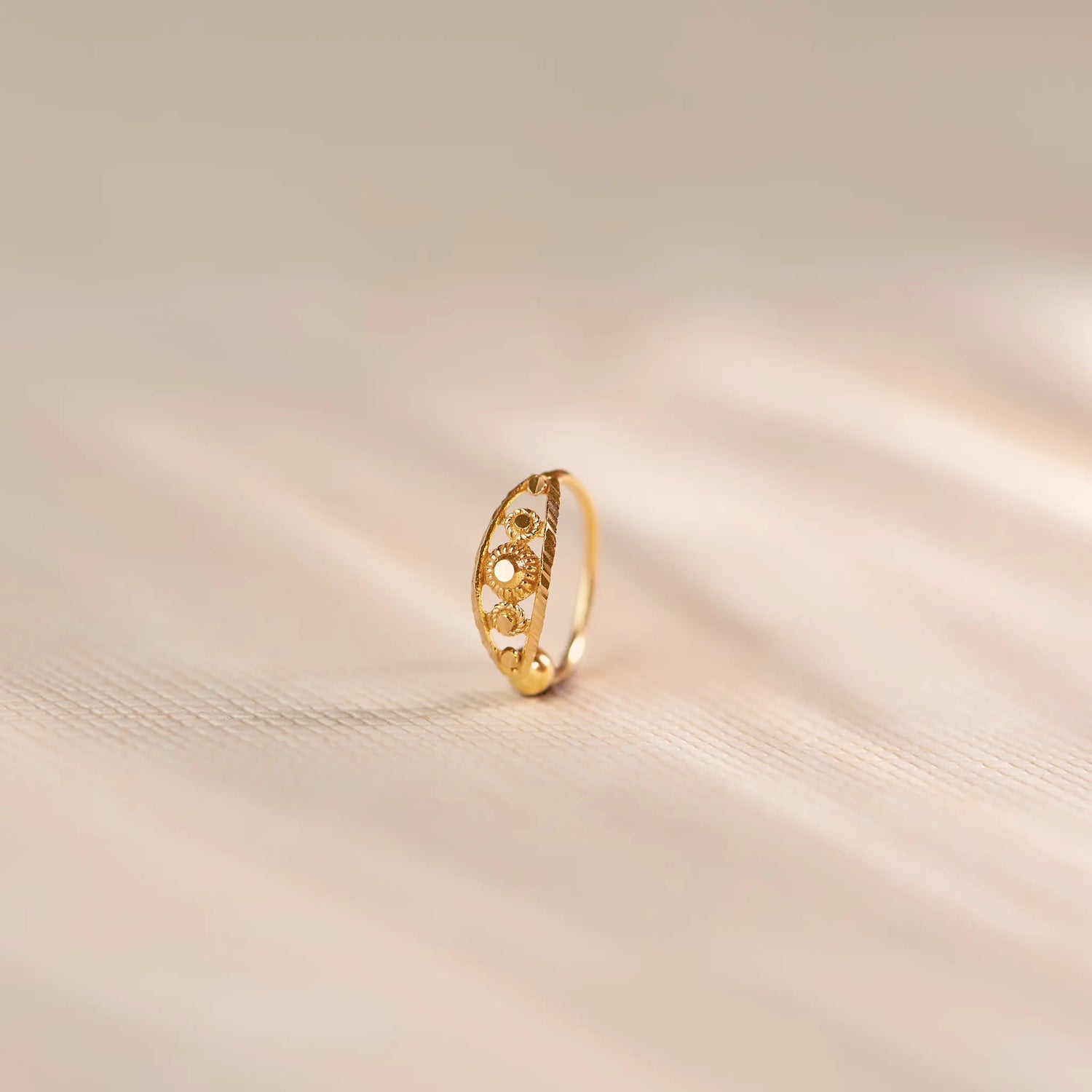 Close up of an 18K gold nose ring with circular cutwork and engraved detailing, standing upright on a soft beige fabric background with gentle light reflections