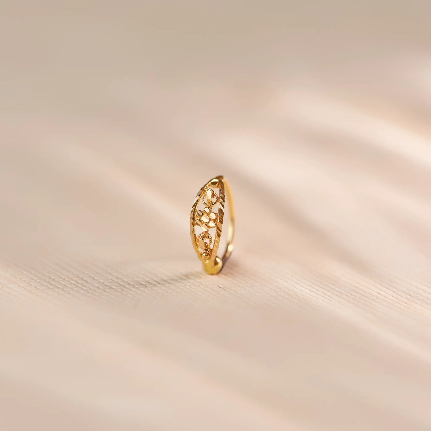 Close up of an 18K gold nose ring with intricate floral cutwork pattern, displayed upright on a soft beige fabric surface with warm natural lighting