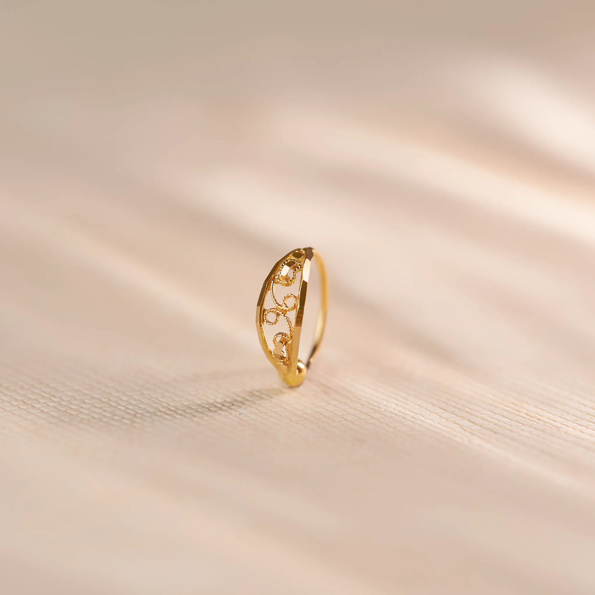 Close up of an intricately cut 18K gold nose ring standing upright on a soft beige fabric background with warm natural light highlighting the delicate pattern