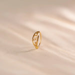 Close up of an intricately cut 18K gold nose ring standing upright on a soft beige fabric background with warm natural light highlighting the delicate pattern