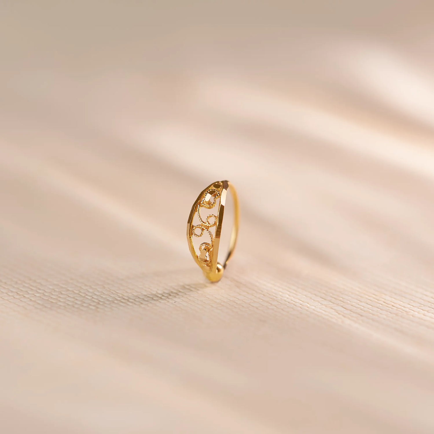 Close up of an intricately cut 18K gold nose ring standing upright on a soft beige fabric background with warm natural light highlighting the delicate pattern