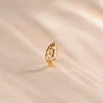 Close up of an ornate 18K gold nose ring with delicate filigree detailing placed upright on a soft beige fabric surface under warm, diffused light