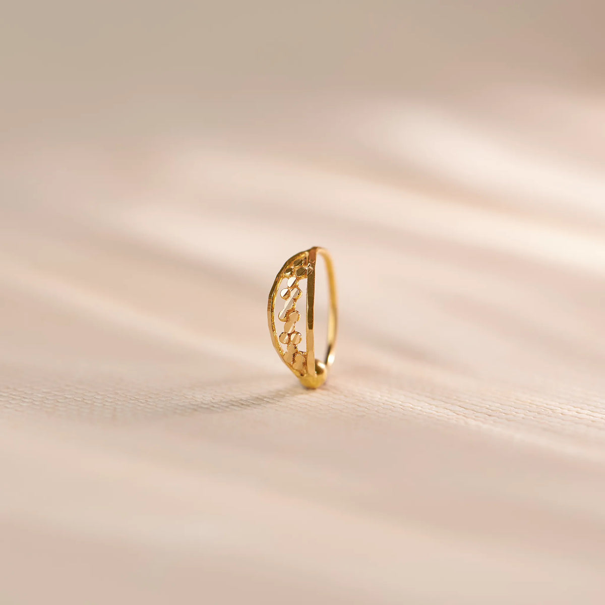 Close up of a slim 18K gold nose ring with intricate cutwork design standing upright on a soft beige textured background with gentle light streaks