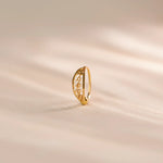 Close up of a slim 18K gold nose ring with intricate cutwork design standing upright on a soft beige textured background with gentle light streaks