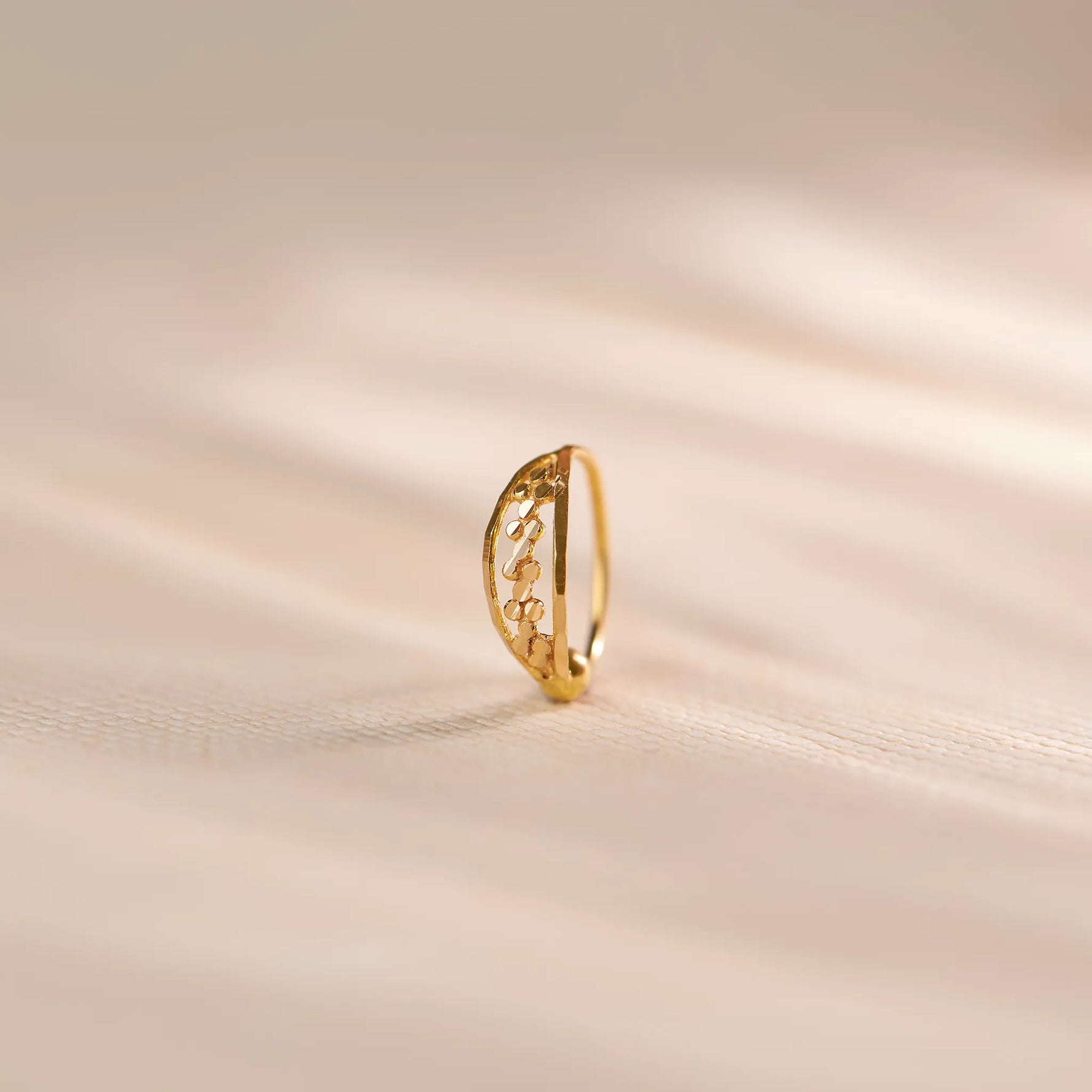 Close up of a slim 18K gold nose ring with intricate cutwork design standing upright on a soft beige textured background with gentle light streaks