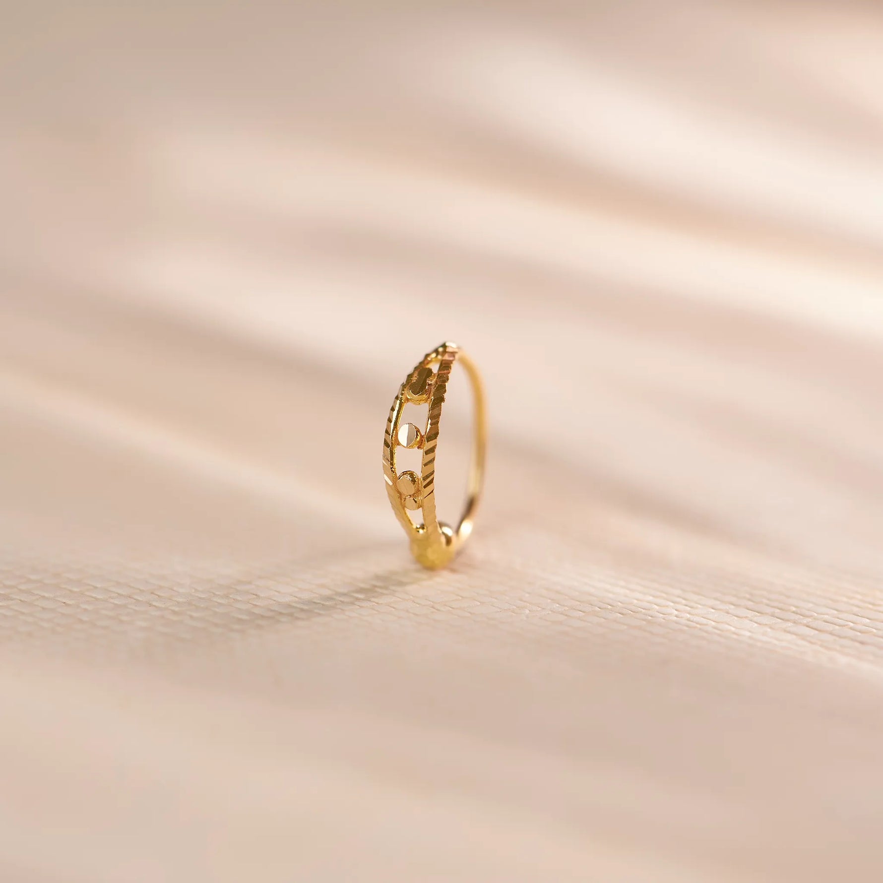 Close up of a delicate 18K gold nose ring with geometric cutwork detailing placed upright on a soft beige surface with subtle flowing light patterns