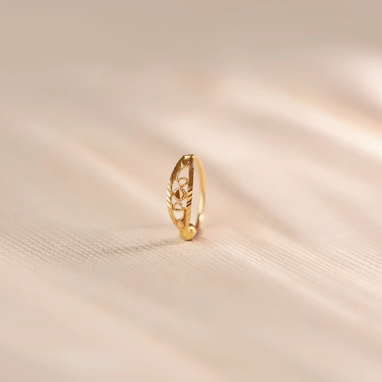 Close up of an 18K gold nose ring with intricate cutwork detailing standing upright on a soft beige fabric background with gentle light reflections
