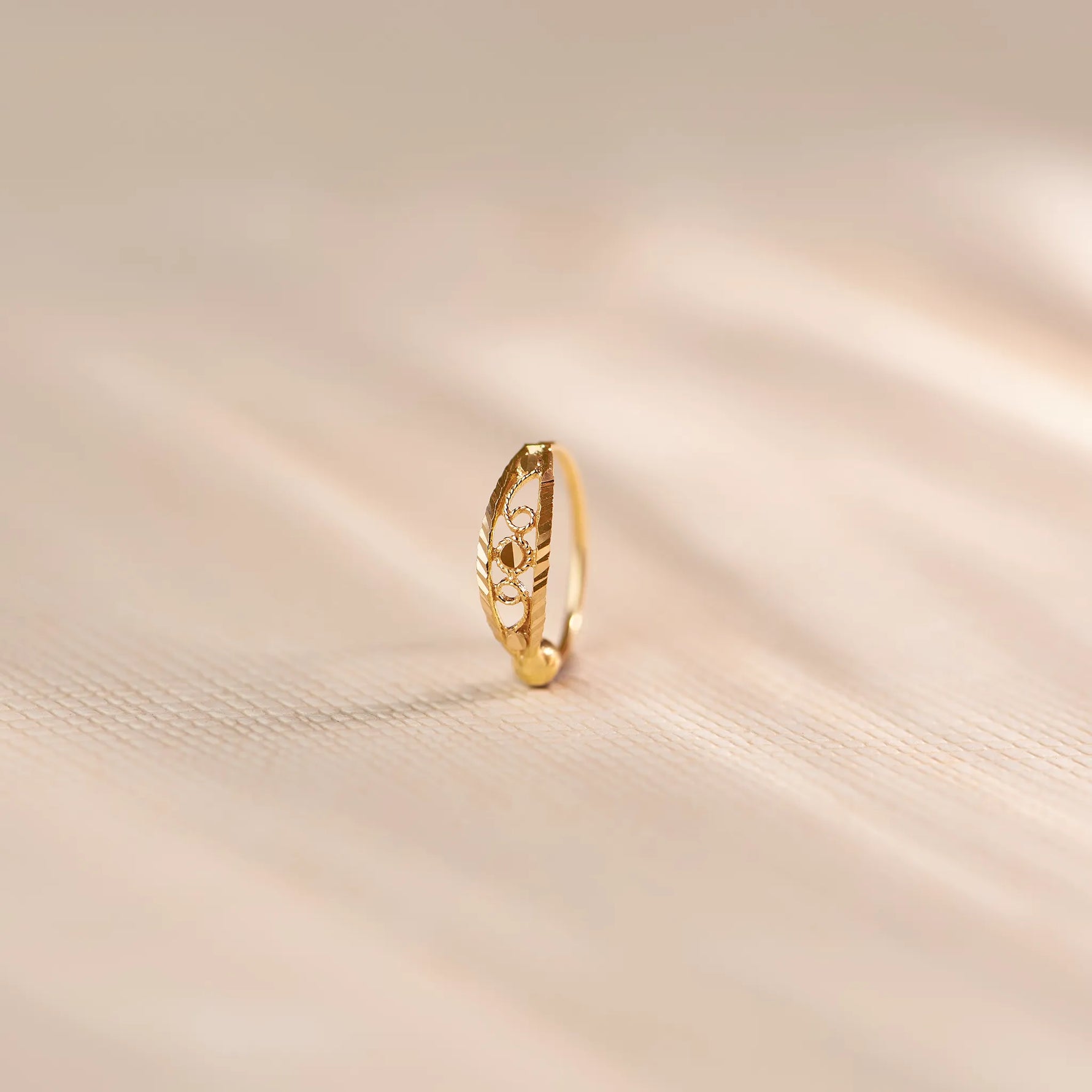 Close up of an 18K gold nose ring with intricate cutwork detailing standing upright on a soft beige fabric background with gentle light reflections