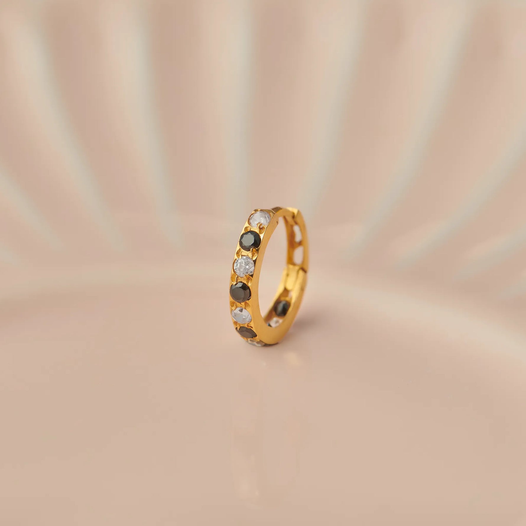 Close up of 18K gold hoop nose ring with black and clear stones placed on a soft beige background highlighting gemstone detailing