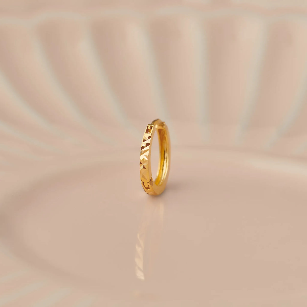 Front view of a slim textured 18K gold hoop nose ring standing upright on a soft beige background with subtle light reflections