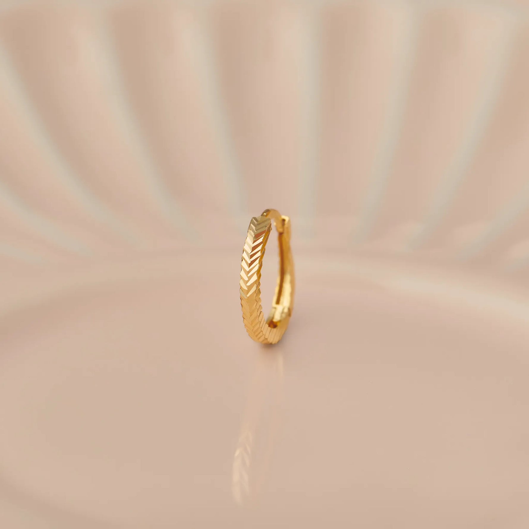 Side view of textured 18K gold hoop nose ring standing upright on a soft beige background with gentle light rays