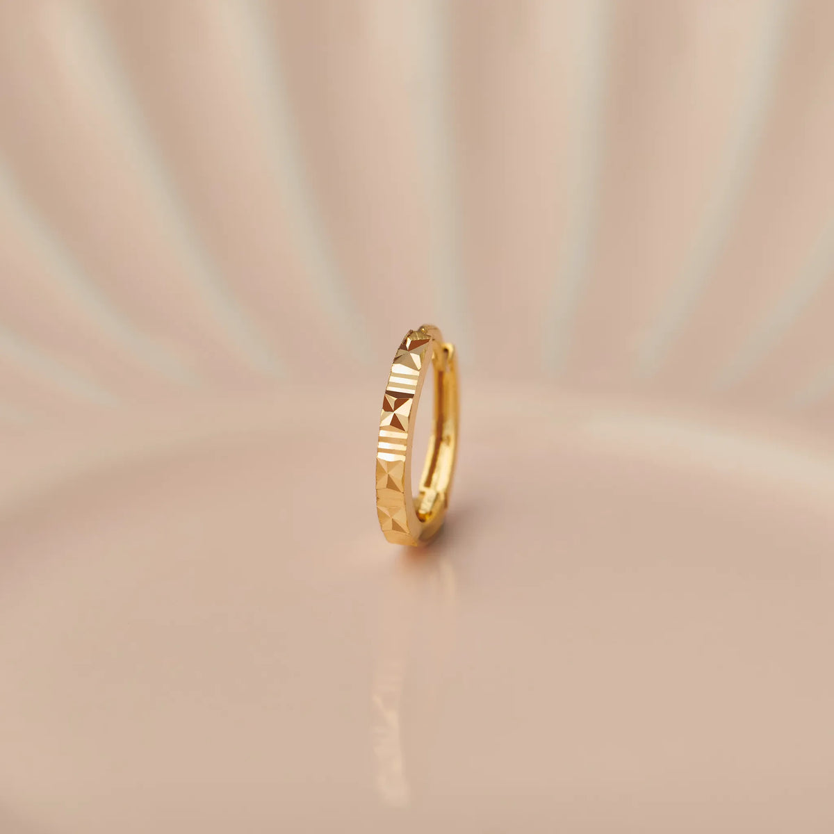 Close up of slim textured gold nose ring placed upright on a soft peach toned surface with gentle studio lighting
