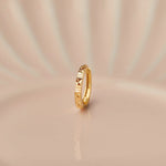 Close up of slim textured gold nose ring placed upright on a soft peach toned surface with gentle studio lighting