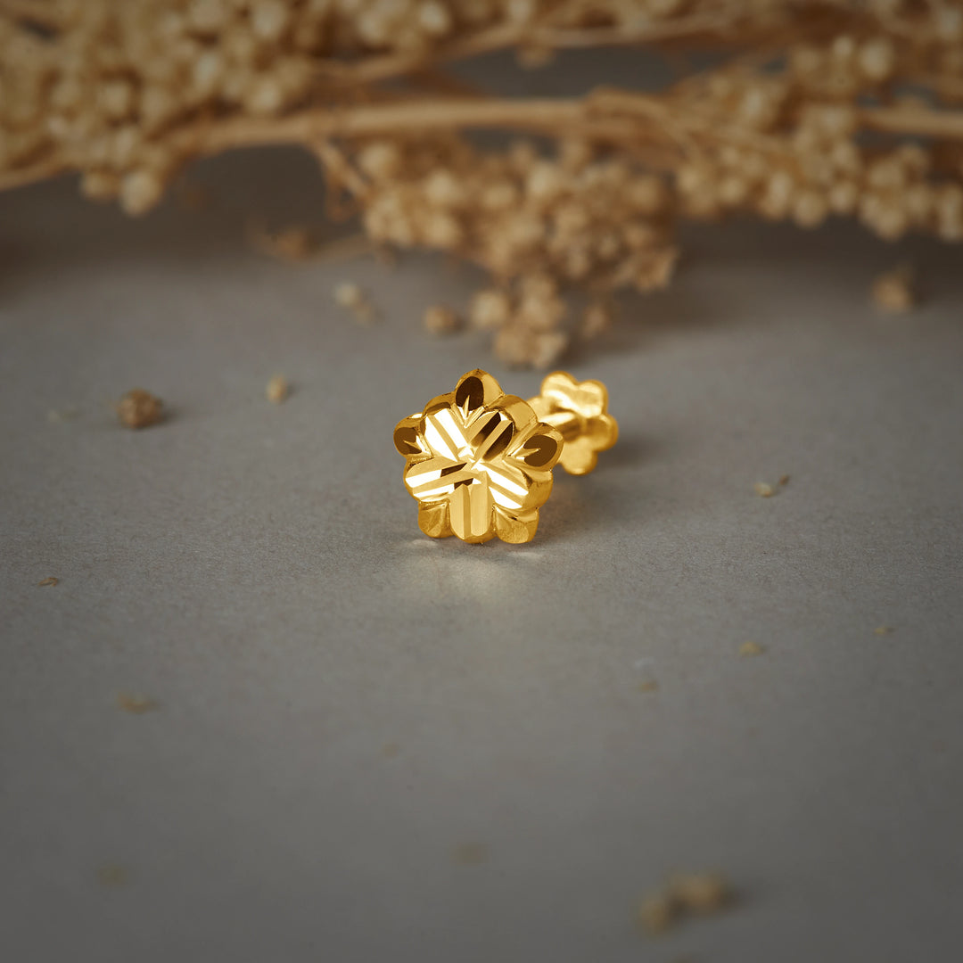 A pair of gold flower-shaped stud earrings with a diamond-cut striped finish, placed on a grey background with dried flowers