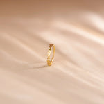 Close up of an 18K gold nose ring with delicate openwork swirl design standing upright on a soft beige textured background with warm light shadows